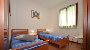 A room with two wooden beds, each with a blue bedspread and a pillow, a small white chair, and a window with green curtains. - Villaggio Azalea (Lignano Sabbiadoro)