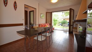 A dining area with a table, chairs, and a sofa. There is a door leading to an outdoor area with a view of greenery. - Villaggio Azalea (Lignano Sabbiadoro)