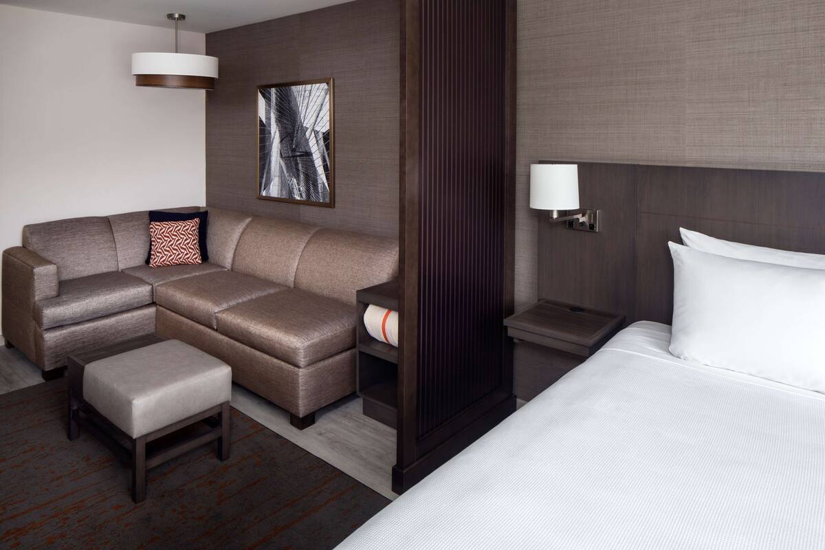 A modern hotel room with a sectional sofa, a small ottoman, a bed with white linens, a nightstand with a lamp, and a framed abstract artwork on the wall.