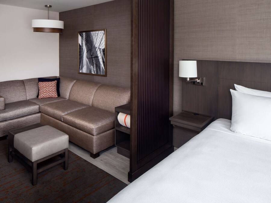 A modern hotel room with a sectional sofa, a small ottoman, a bed with white linens, a nightstand with a lamp, and a framed abstract artwork on the wall.
