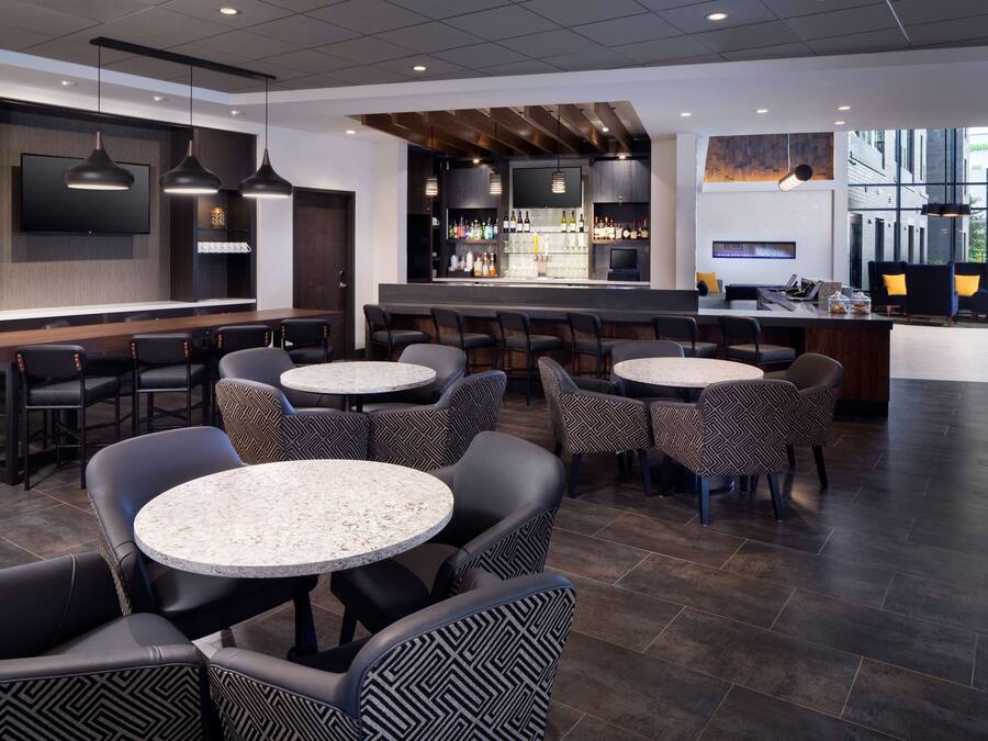 A modern bar area with round tables and chairs, a bar counter with a variety of bottles, and a large screen mounted on the wall.