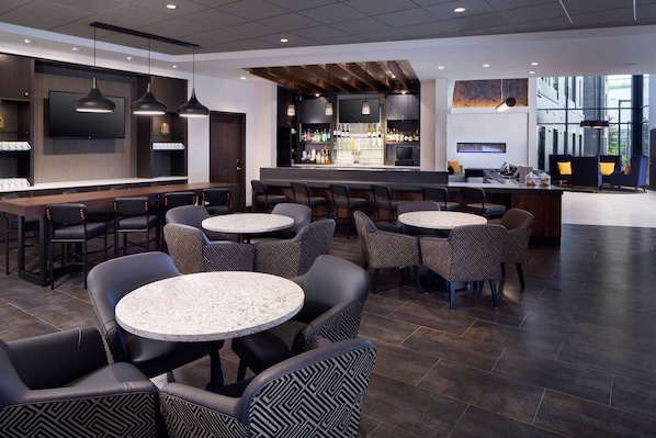 A modern bar area with round tables and chairs, a bar counter with a variety of bottles, and a large screen mounted on the wall.