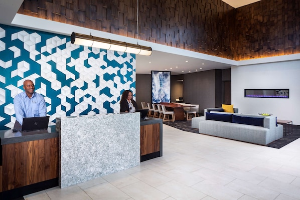 A reception area with a geometric patterned wall, a reception desk, a sofa, and a dining area with chairs and a table. - Hyatt Place Poughkeepsie / Hudson Valley (Poughkeepsie)