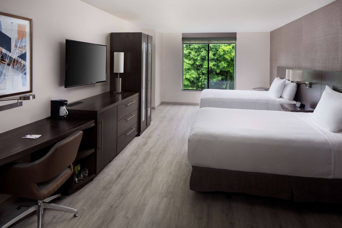 A hotel room with a desk, two beds, a TV, and a window with a view of greenery.
