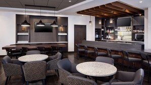 Zona de bar moderna con mesas redondas y sillas estampadas, una barra con televisor y estanterías con botellas.