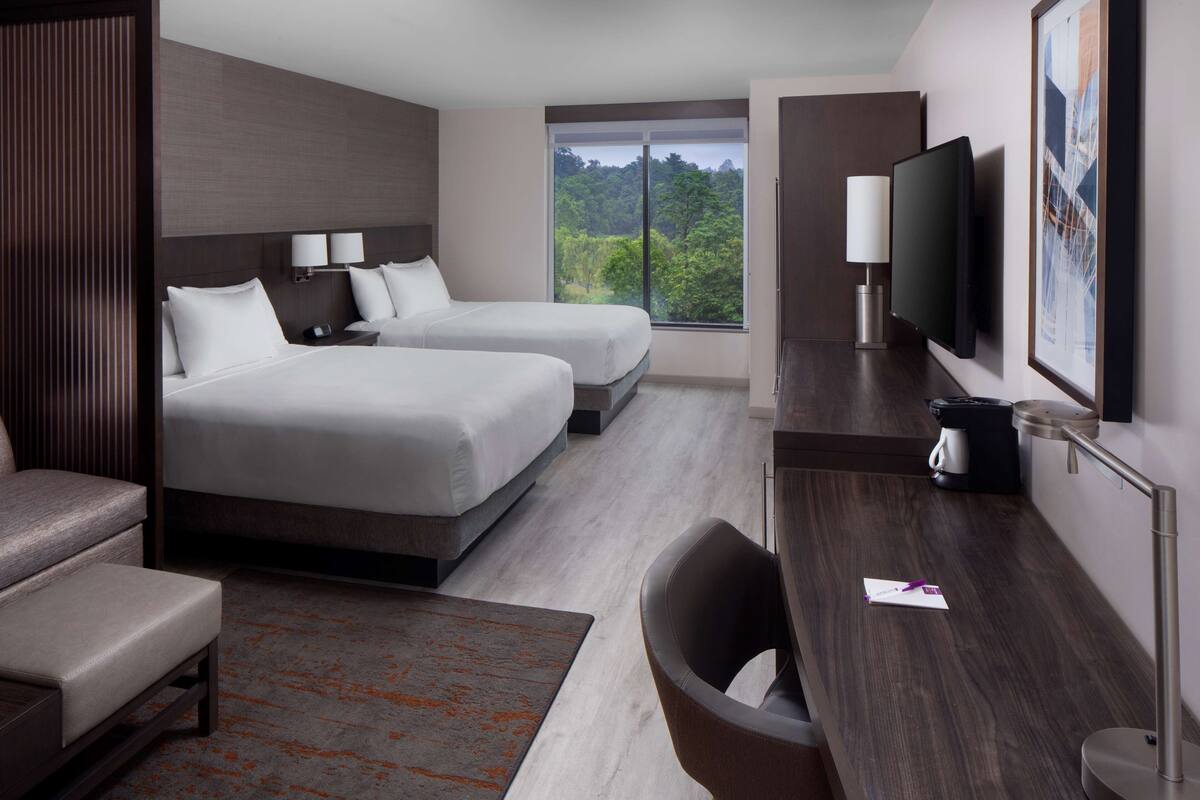 A modern hotel room with two beds, a desk, a chair, a TV, and a large window offering a view of trees.