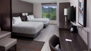 A modern hotel room with two beds, a desk, a chair, a TV, and a large window offering a view of trees.