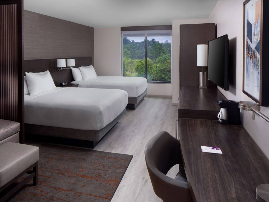 A modern hotel room with two beds, a desk, a chair, a TV, and a large window offering a view of trees.