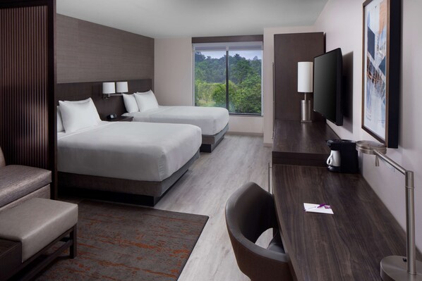 A modern hotel room with two beds, a desk, a chair, a TV, and a large window offering a view of trees.