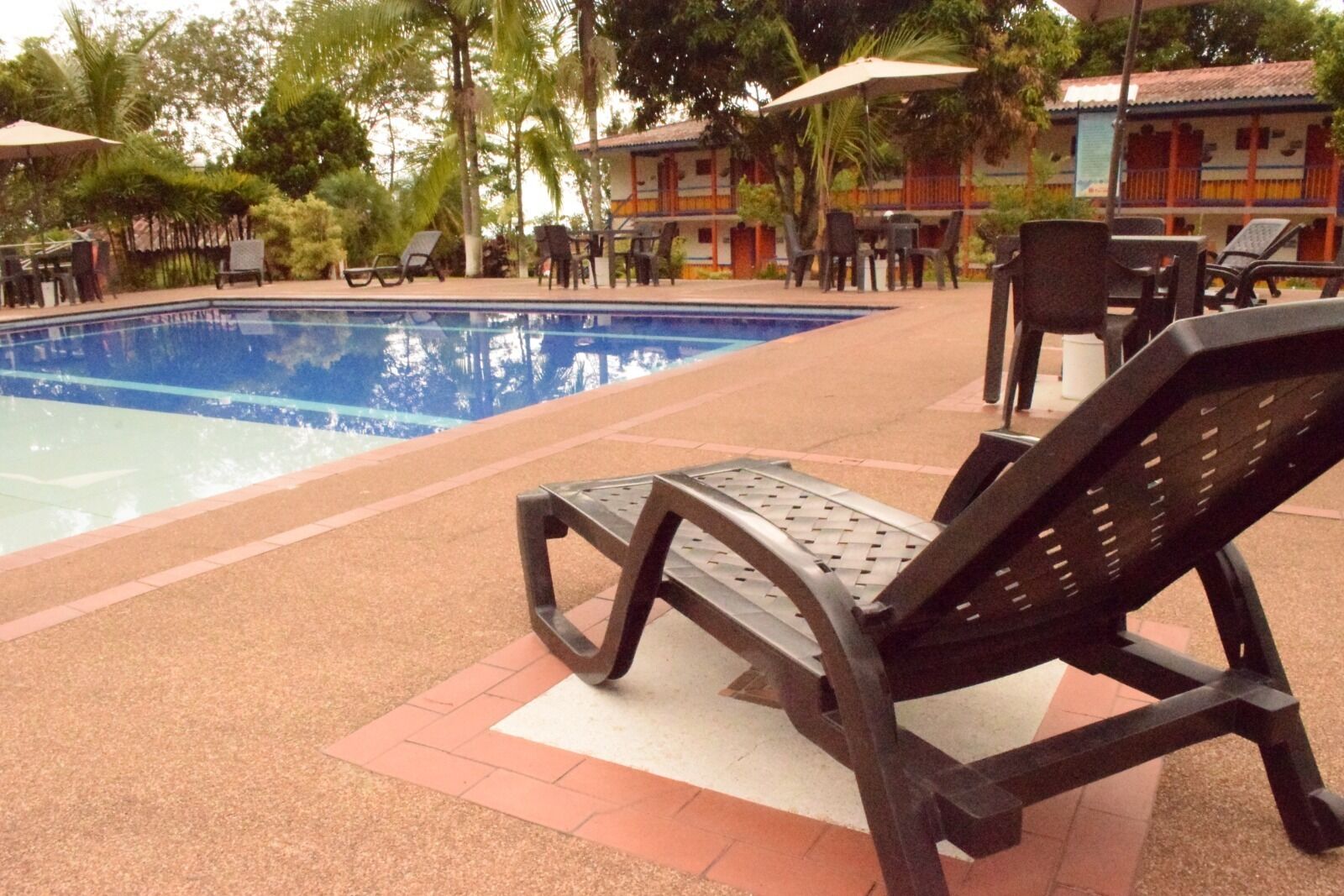 Zona de piscina con sillas de descanso y sombrillas, rodeada por un edificio con tejado de baldosas y una piscina.