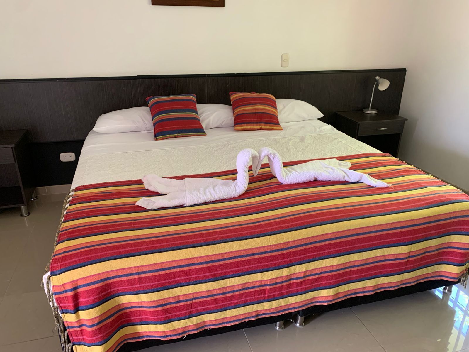 Hotelkamer met een groot bed, een gestreept beddegoed, twee kussens, een nachtkastje, een lamp en een stopcontact.