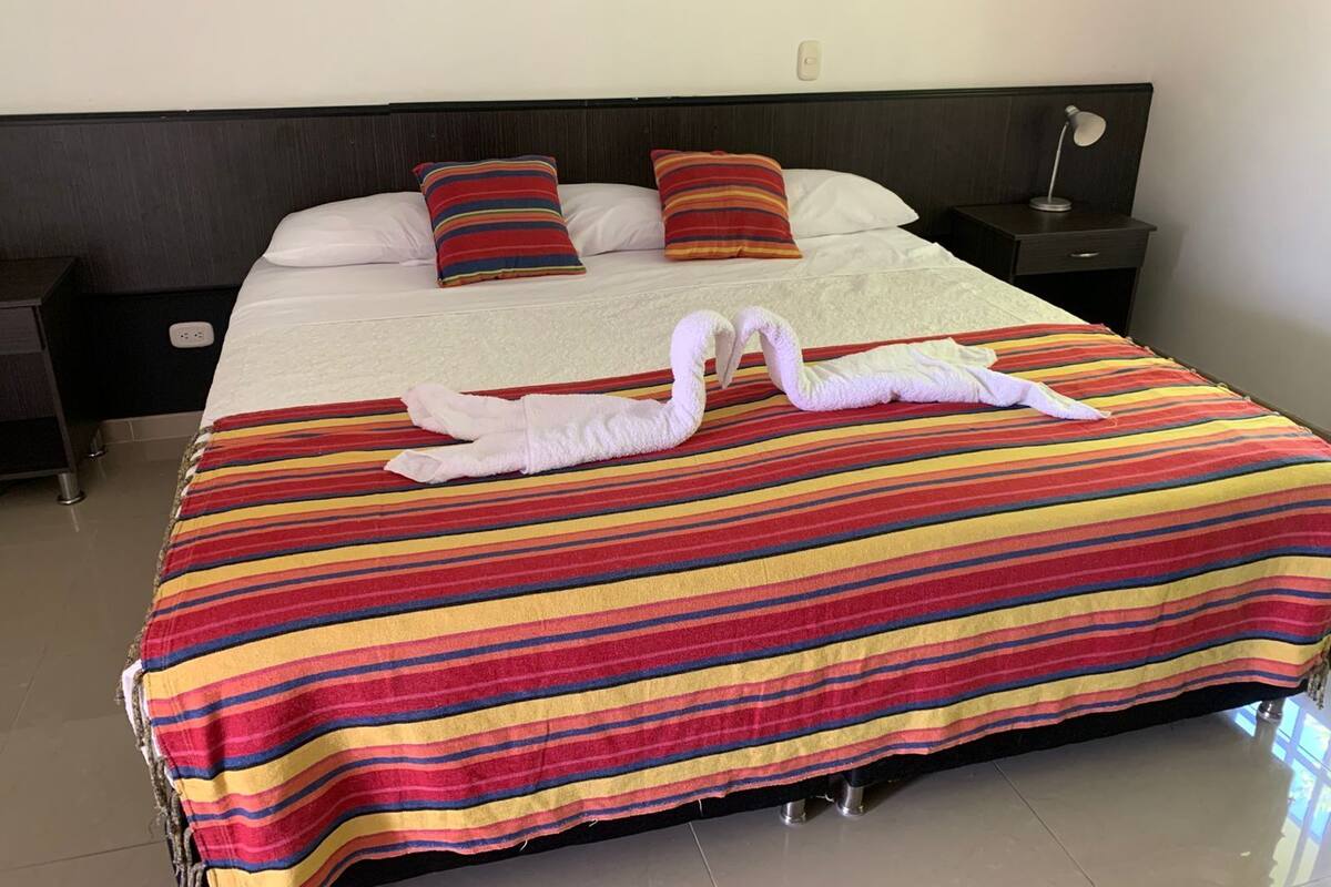 A hotel room with a large bed, striped bedspread, two pillows, a nightstand, a lamp, and a wall socket.