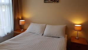 A hotel room with a bed, two bedside lamps, a window with curtains, and a cityscape painting on the wall. - Royal Windsor Holiday Apartments (Blackpool)