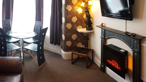 A cozy living room with a fireplace, a flat-screen TV mounted on the wall, a round glass table with chairs, and a decorative wall with floral wallpaper. - Royal Windsor Holiday Apartments (Blackpool)