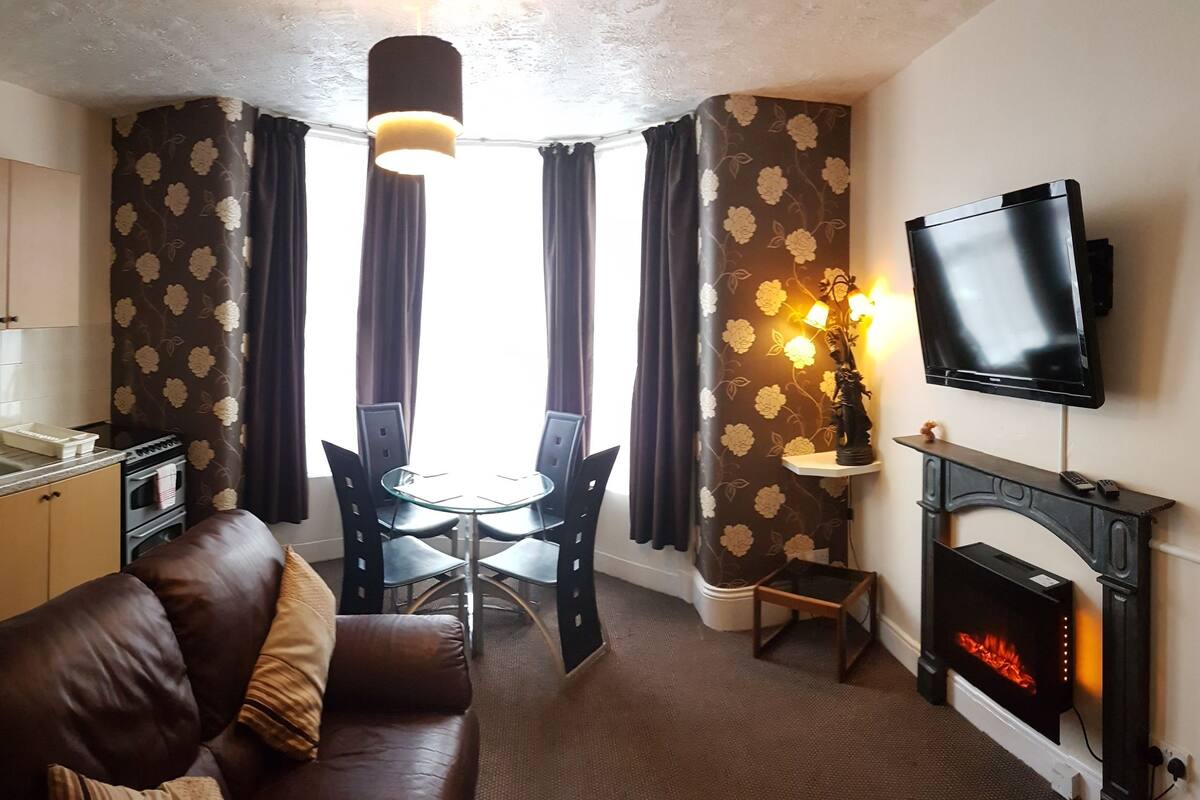 A compact living space with a kitchenette, a dining area, a sofa, a fireplace, and a TV mounted on the wall.