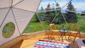 Habitación en forma de domo geodésico, con piso de madera, una pequeña mesa redonda con un florero, dos sillas de madera y vistas a un bosque a través de amplios ventanales.