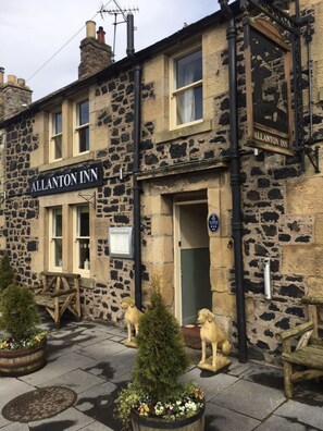 Step into the charming entrance of The Allanton Inn, where a welcoming bench invites you to relax amidst a beautifully maintained garden with potted plants and a small tree.