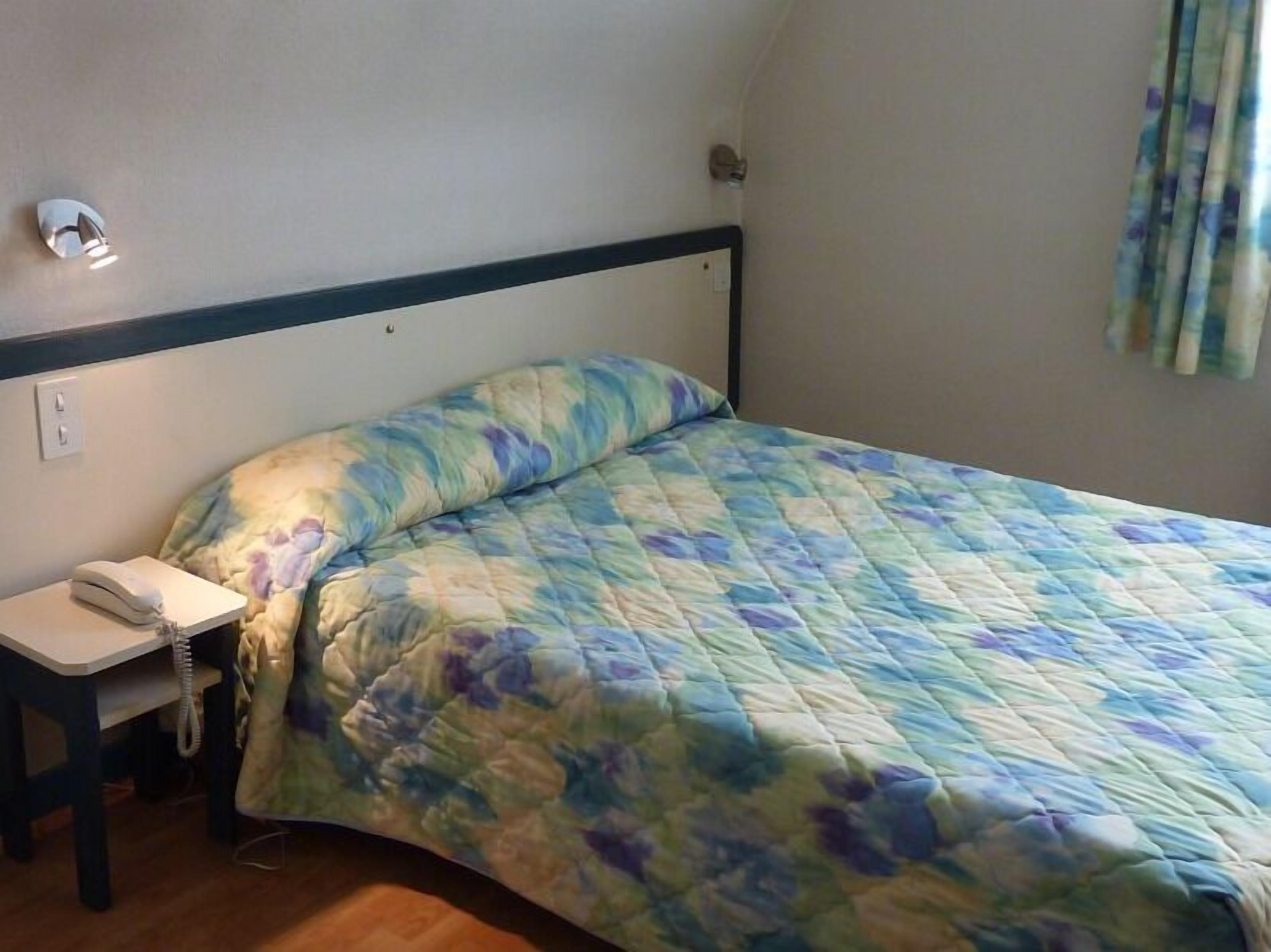A single bed with a floral quilt, a bedside table with a phone, and a wall-mounted light fixture.