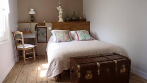 Une chambre avec un plancher en bois, un lit agrémenté d’oreillers à motifs floraux, une chaise en bois, un bureau et une grande valise marron.