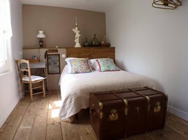 A bedroom with a wooden floor, a bed with floral pillows, a wooden chair, a desk, and a large brown suitcase.