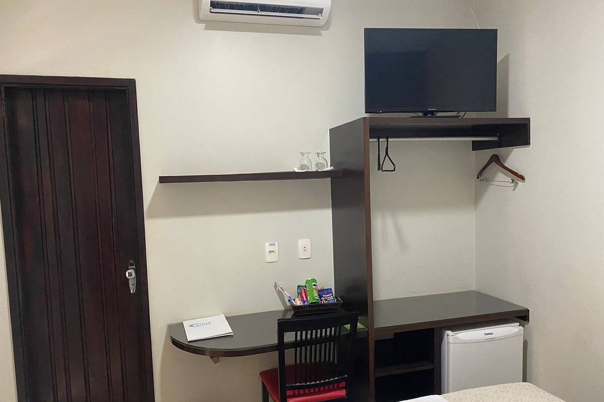 Ein kleines Hotelzimmer mit einem Bett, einem Schreibtisch mit Stuhl, einem an einer Wandhalterung befestigten Fernseher, einem kleinen Kühlschrank und einer Tür.