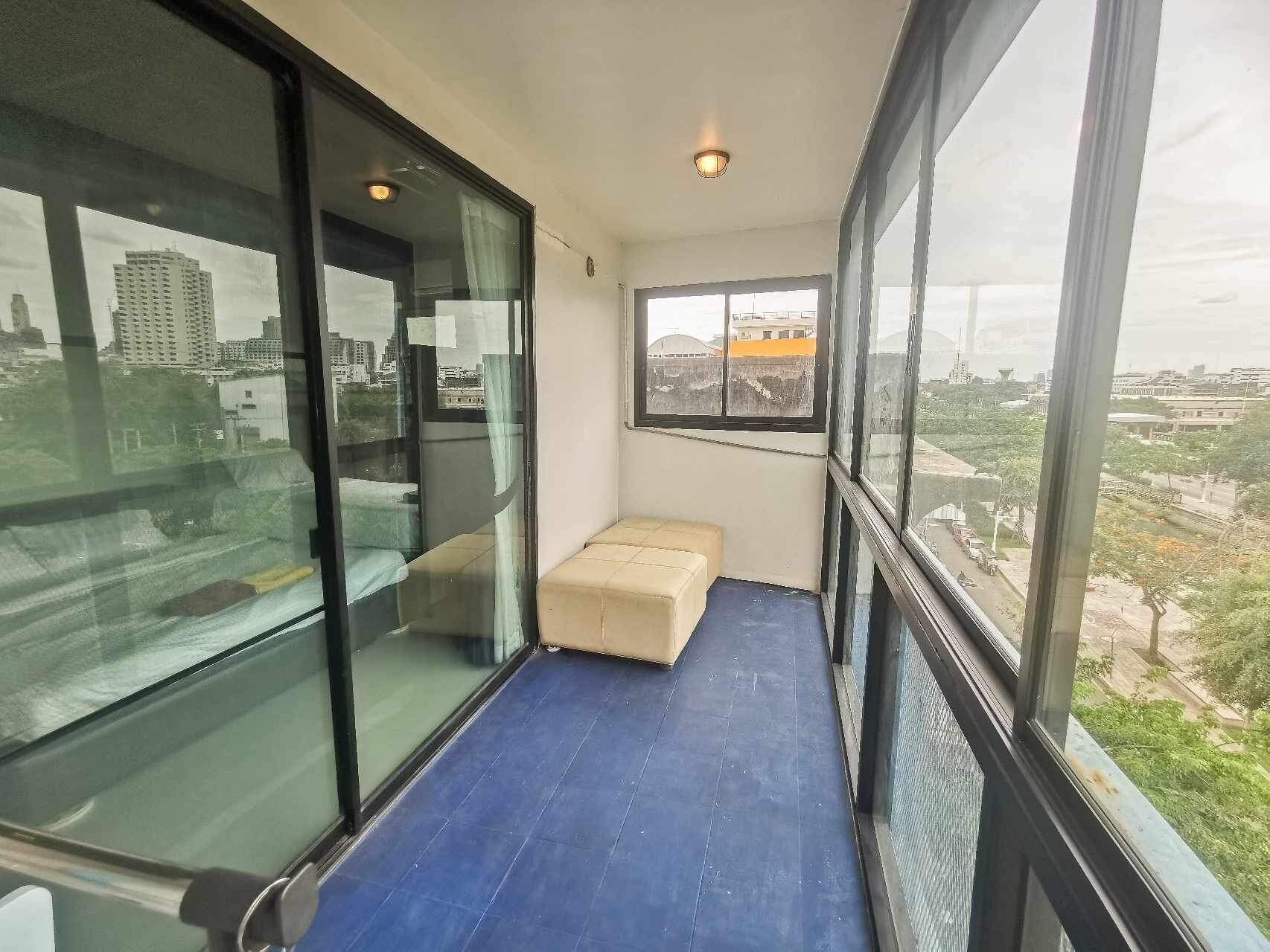 A balcony with a blue floor, a beige bench, and a view of a cityscape through large windows.