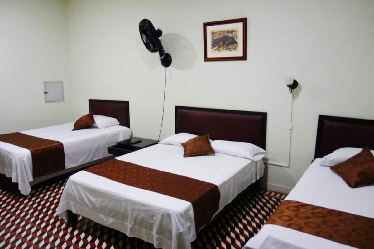A hotel room with three single beds, each with a brown headboard and white bedding. There is a black fan on the wall, a framed picture, and a whiteboard on the wall.