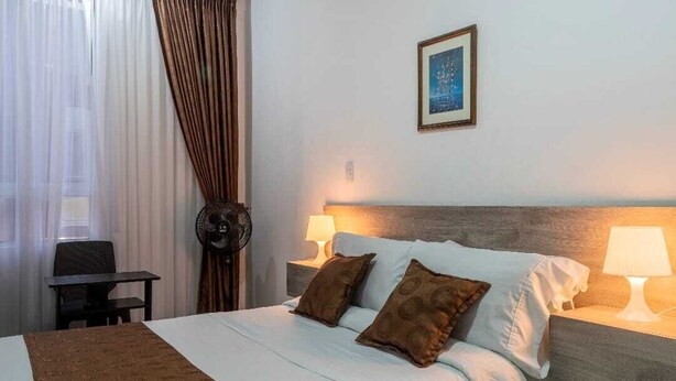 Quarto de hotel com cama, duas luminárias de cabeceira, uma moldura na parede e uma janela com cortinas.