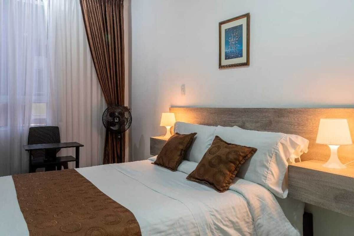 A hotel room with a bed, two bedside lamps, a framed picture on the wall, and a window with curtains.