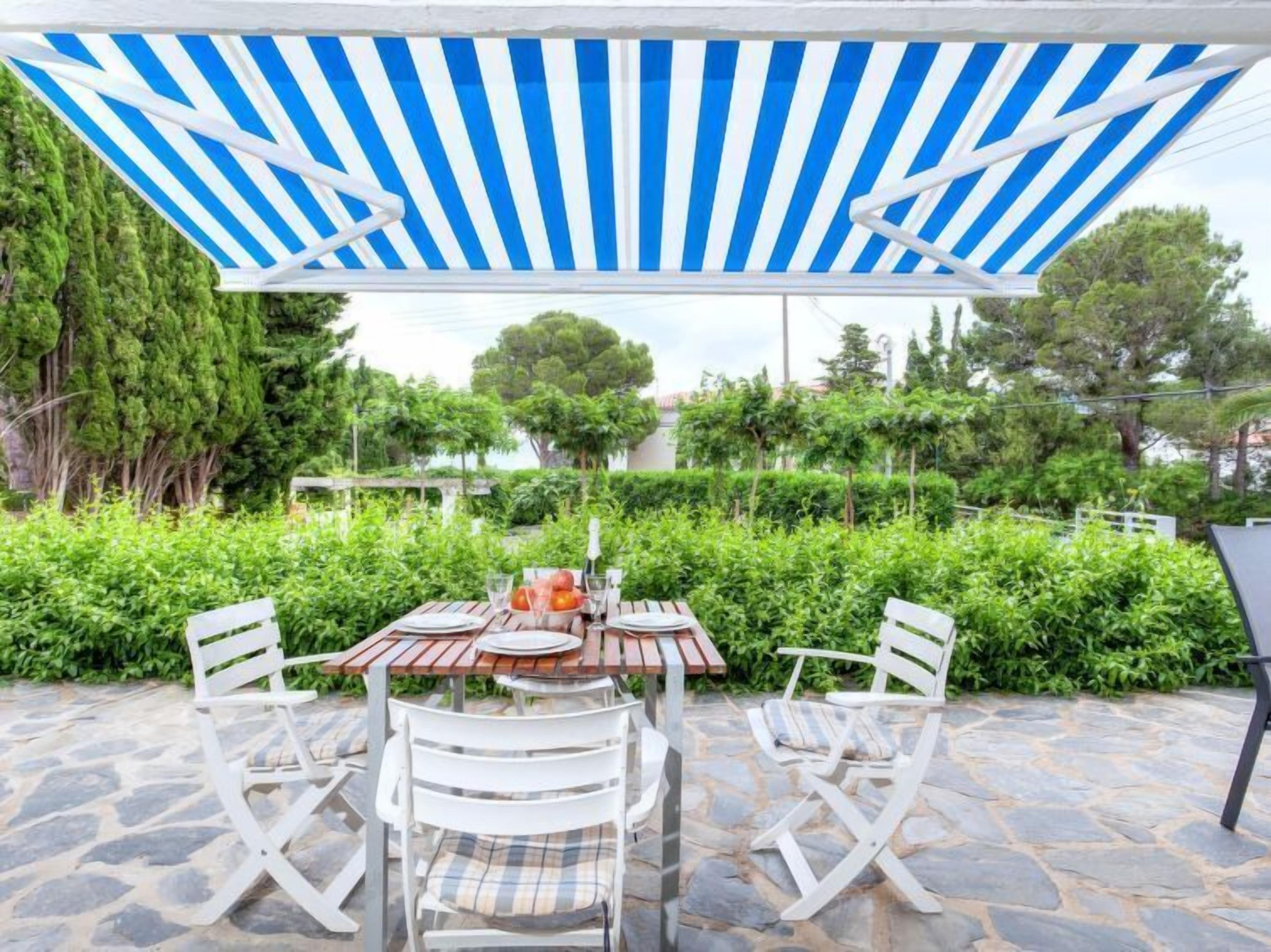 Un patio al aire libre con un toldo a rayas, suelo de piedra y sillas plegables blancas.