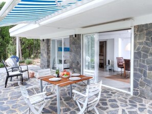 Um pátio com uma mesa posta para refeições, piso de pedra e uma porta de vidro que dá para um espaço interno.
