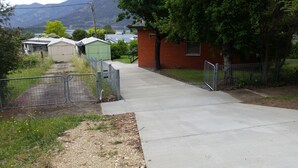 Un chemin en béton mène à une zone délimitée par une grille, où se trouvent un petit bâtiment en briques et un abri de jardin vert.