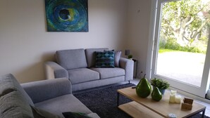 A modern living room with a grey sofa, a wooden coffee table, and a large window offering a view of greenery outside. - Villa Oostduinkerke, 400m sea+dunes,quiet+green+much light, 6p+baby,3bed+2bath (Koksijde)