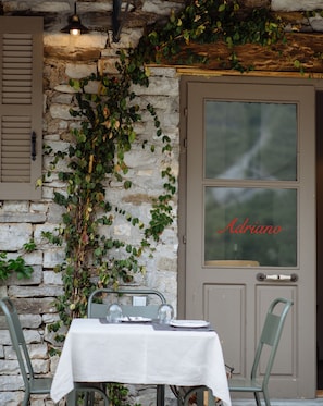 A rustic stone wall with a wooden door labeled "Adriano," a table with a white tablecloth, and metal chairs.