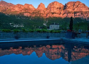 A pool reflecting mountainous terrain, a stone building, and lush greenery. - Avragonio (Zagori)