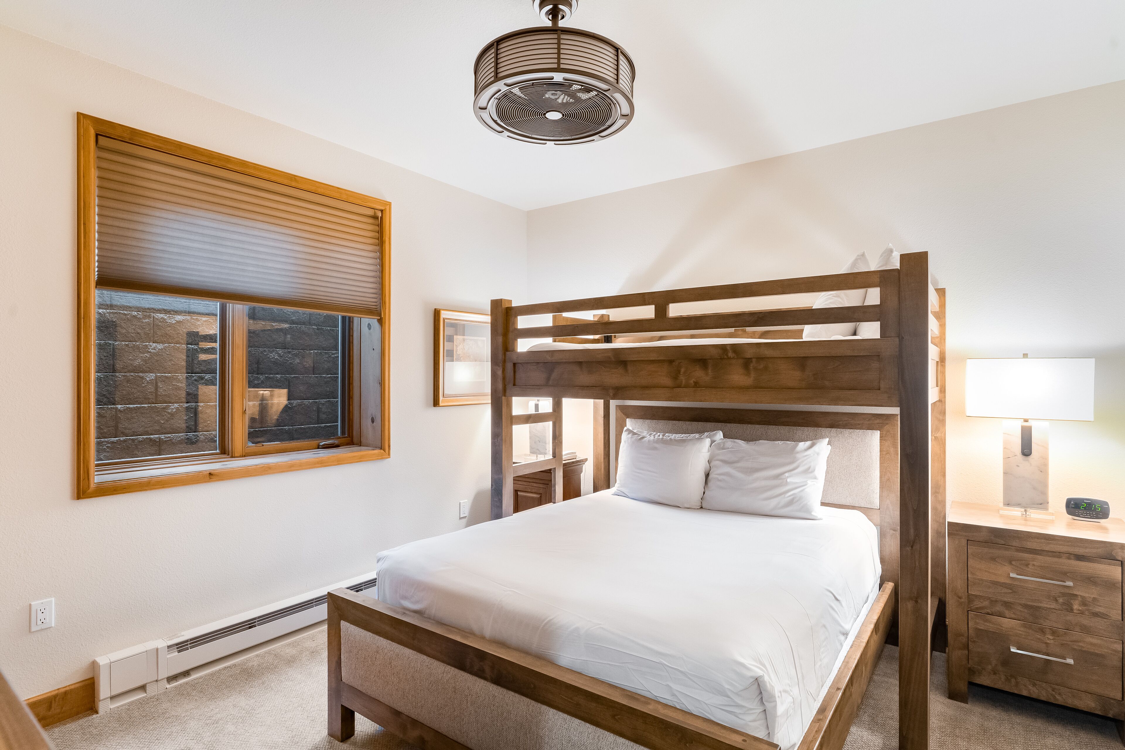 A bedroom with a bunk bed, a window with blinds, a bedside table with a lamp, and a ceiling fan.