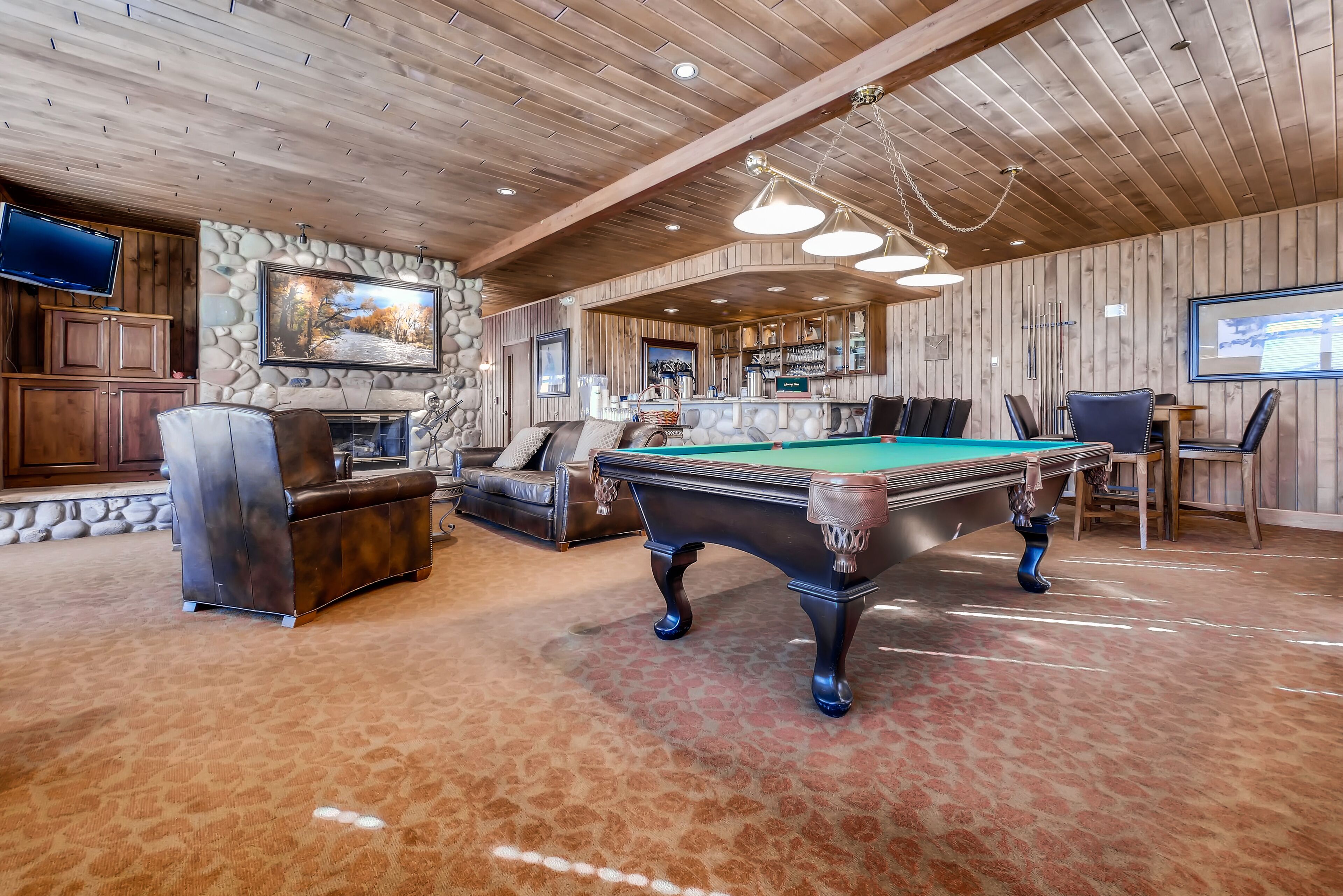 Une table de billard dans une pièce aux murs et au plafond en bois, avec une cheminée en pierre et un coin bar.