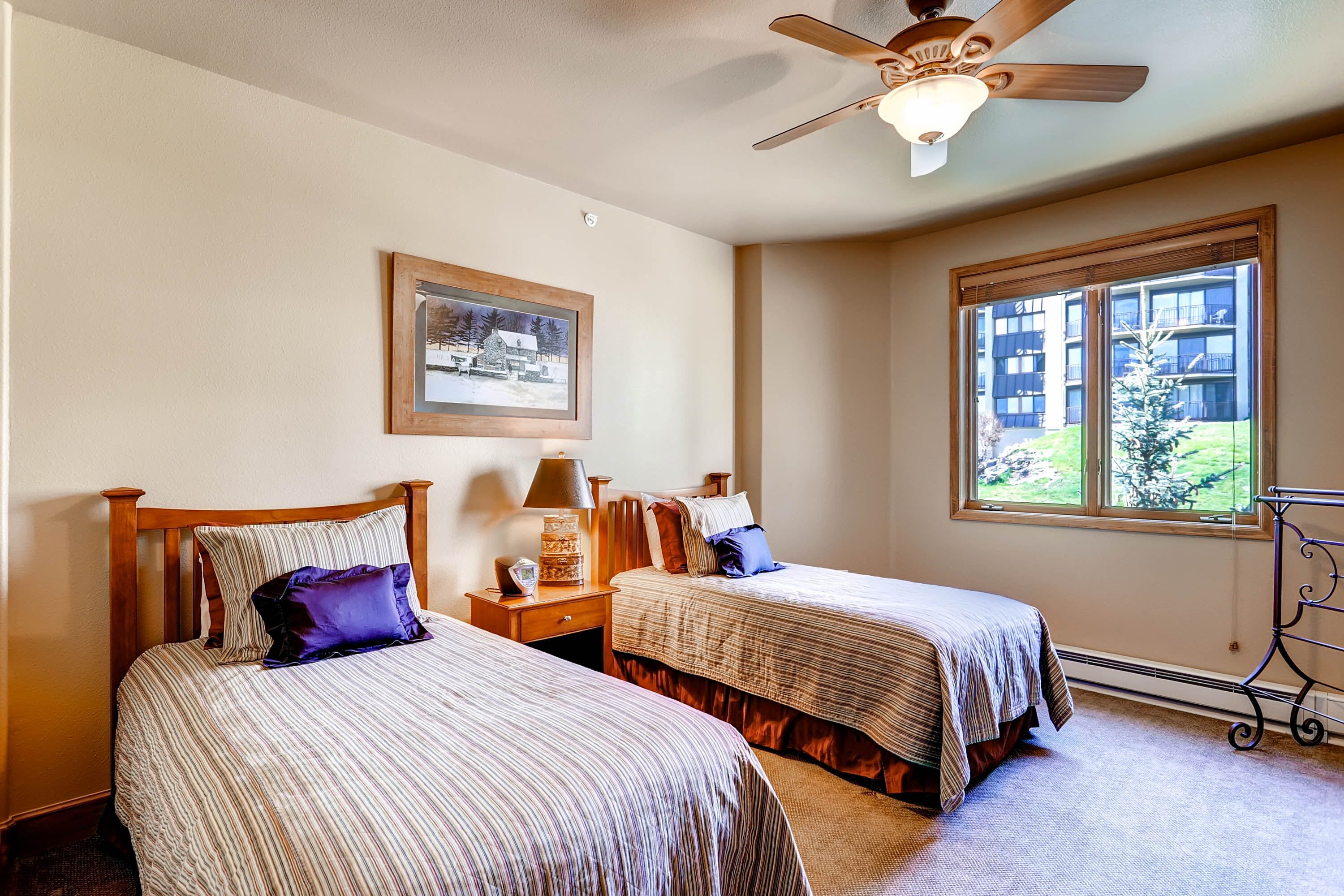 A hotel room with two beds, a ceiling fan, a window with a view of a building, and a framed picture on the wall.