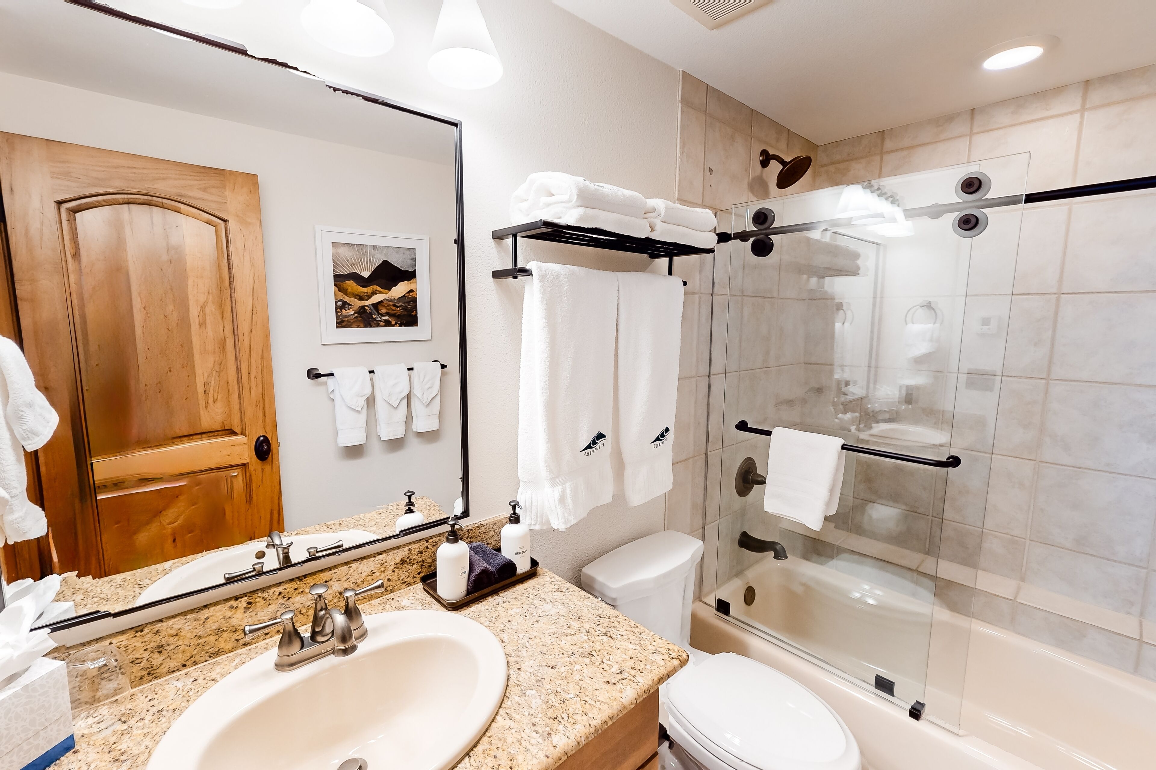 Une salle de bain moderne avec un comptoir en granit, un grand miroir, une porte en bois, une douche en verre et des toilettes.