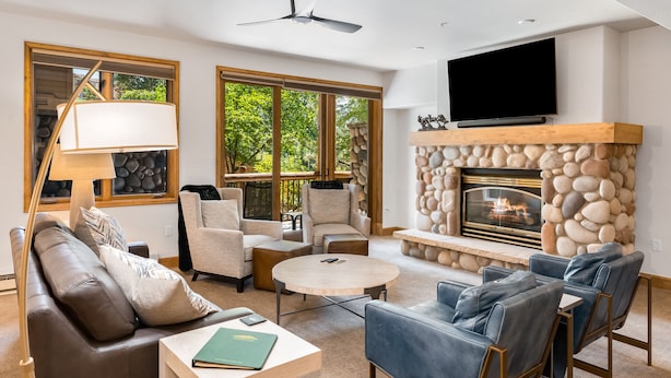 A modern living room with a stone fireplace, comfortable seating, a flat-screen TV, and large windows offering a view of the outdoors.