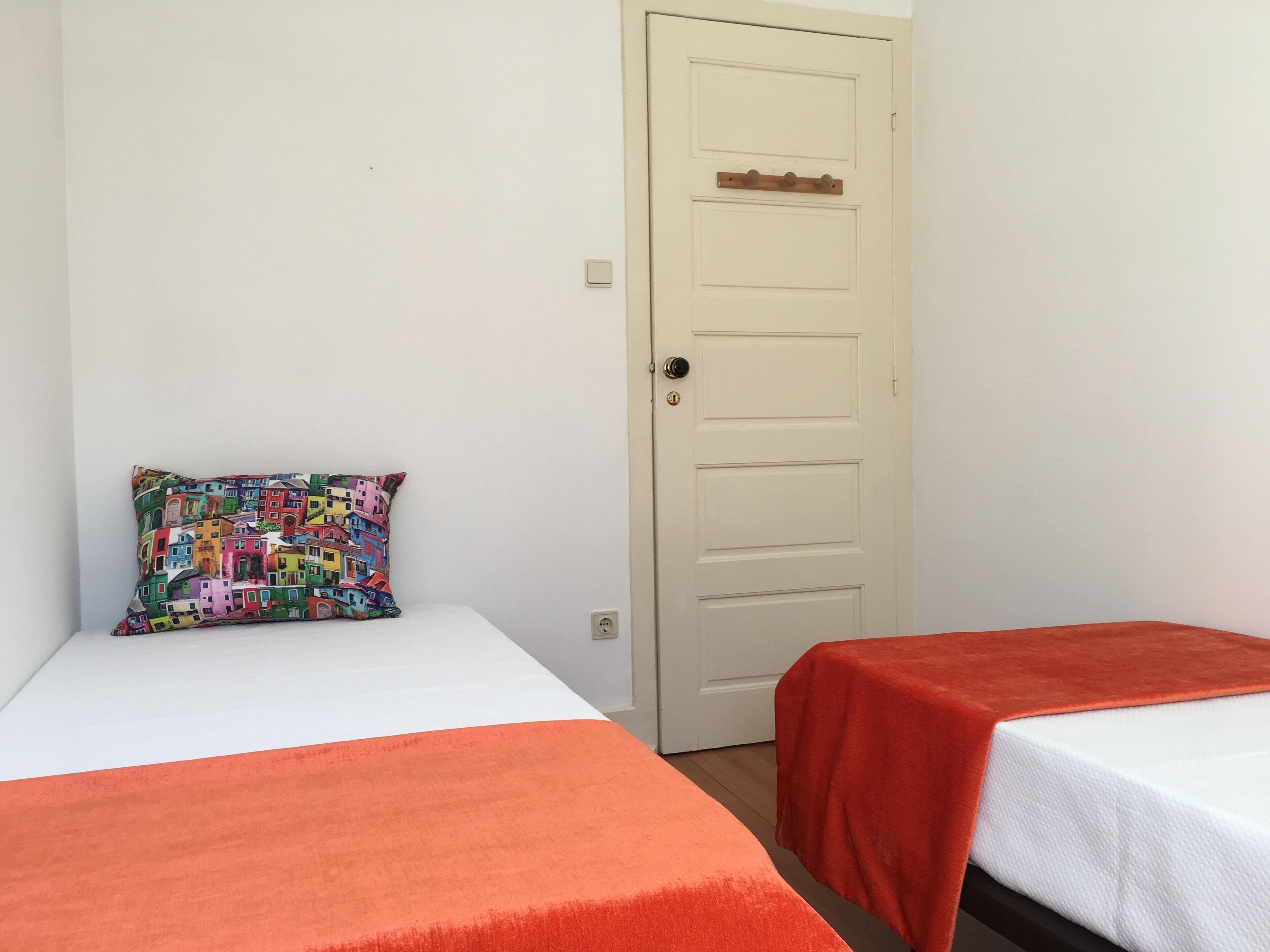 Una habitació amb dos llits, cadascun cobert amb un full blanc i una manta taronja. Un coixí de colors reposa sobre un dels llits. Al fons, es veu una porta blanca amb un petit panell de fusta.