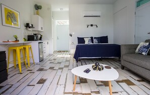 Een compact leefruimte met een kitchenette, een bed met donkerblauwe beddengoed, een grijze bank en een ronde witte tafel met een afstandsbediening en decoratieve objecten.