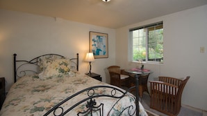 A bedroom with a bed, a bedside table, a lamp, a chair, a small table, and a window with a view of greenery. - River View Garden Resort (Monte Rio)