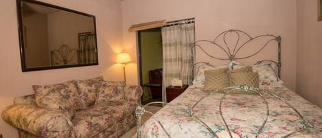 A bedroom with a floral-patterned bed, a matching sofa, a bedside table with a lamp, and a large mirror on the wall.
