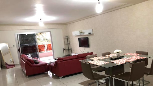 A modern living room with a glass dining table, red sofas, and a TV mounted on the wall. - Résidence italien2  T3 (Abidjan)