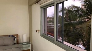 A bedroom with a bed, bedside table, lamp, and a window with a view of palm trees and buildings. - Résidence italien2  T3 (Abidjan)