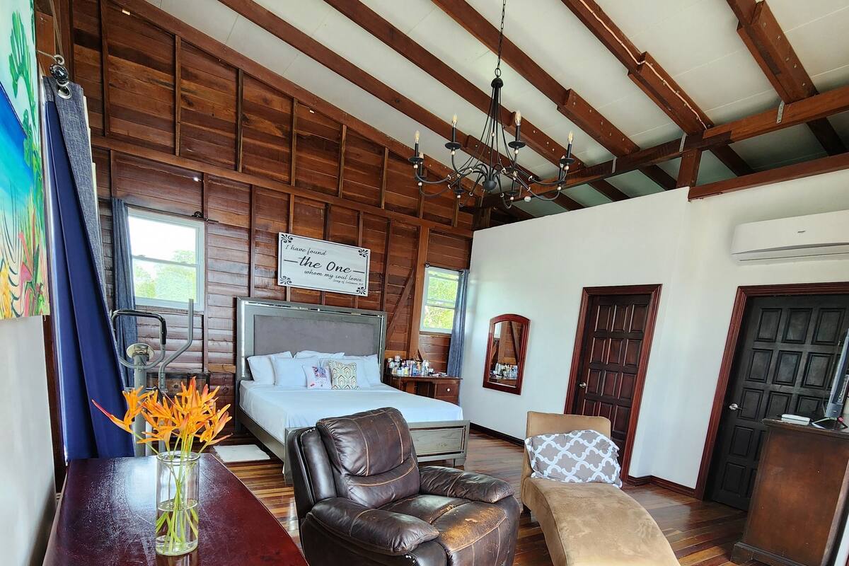 Una habitación amplia con techo de madera, una cama grande con cabecera, un sillón de cuero reclinable, una mesa de comedor de madera con un jarrón de flores y un espejo grande en la pared.