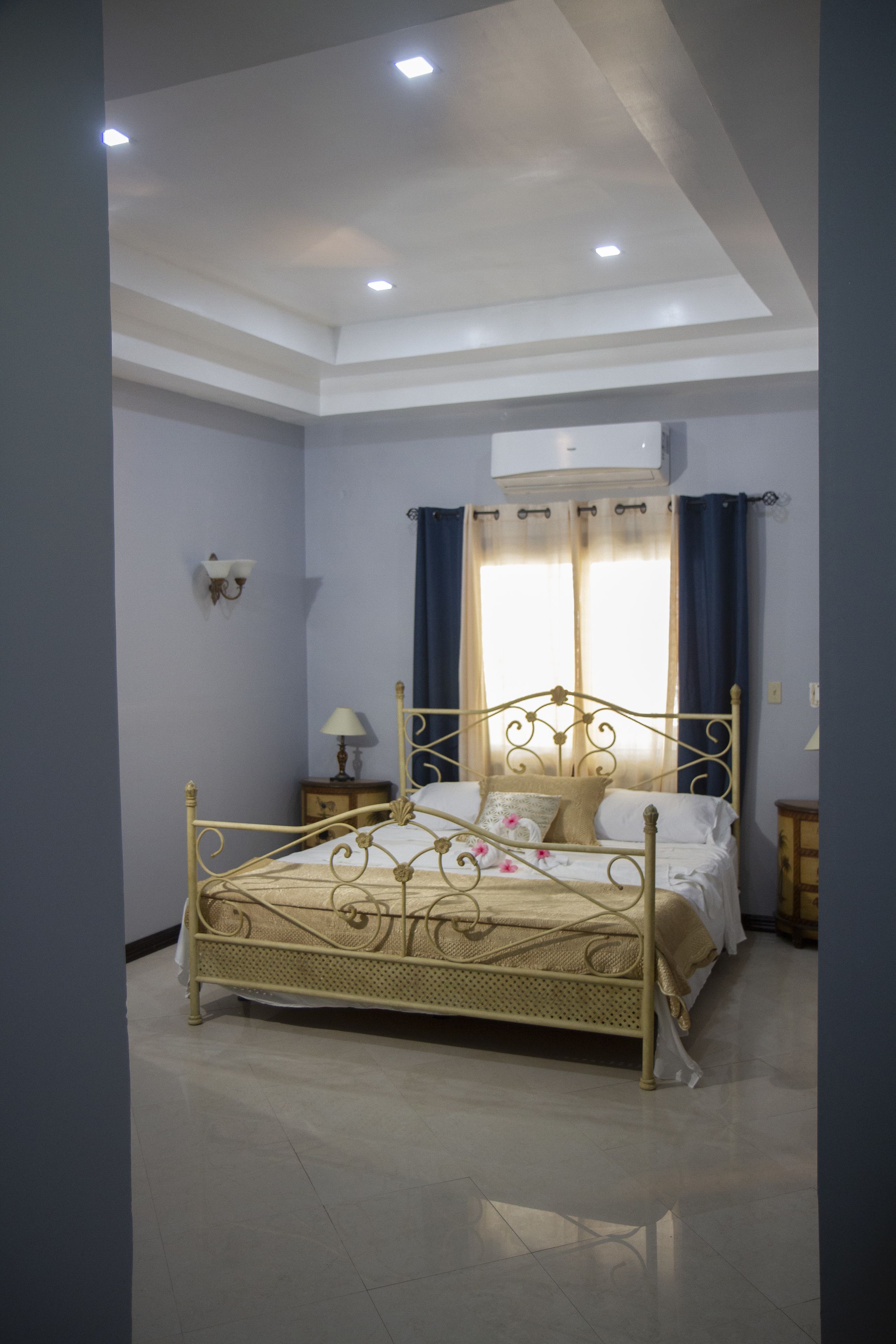Une chambre à coucher avec un lit en métal, une table de chevet avec une lampe, une fenêtre avec des rideaux et un appareil de climatisation.