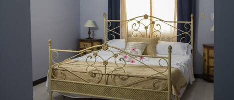 Un dormitorio con estructura de cama de metal, una mesita de noche con lámpara, una ventana con cortinas y un equipo de aire acondicionado.