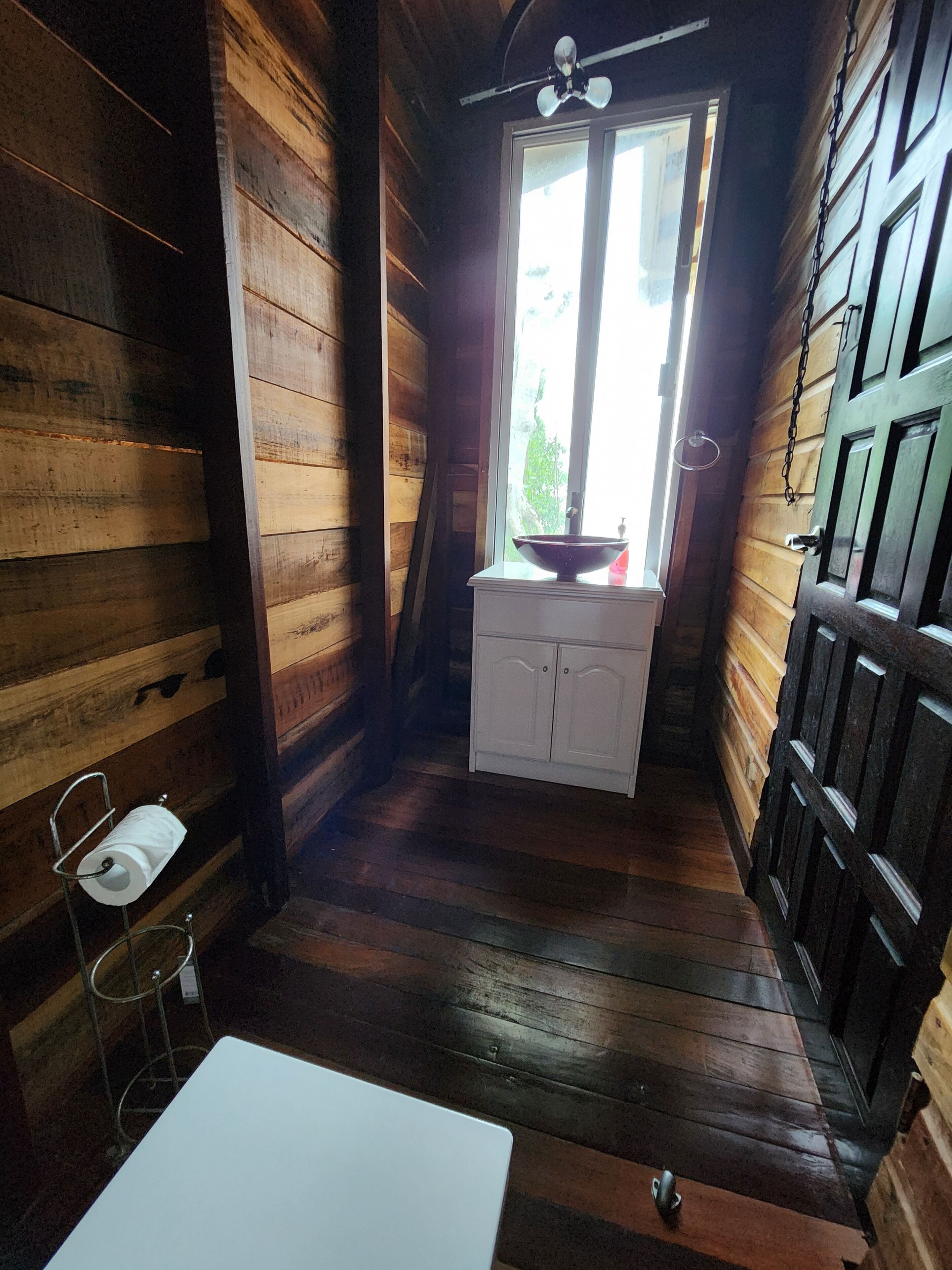 A narrow wooden cabin with a small white cabinet, a white sink, and a metal rack holding a roll of toilet paper.